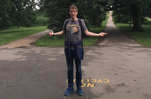 Man with rucksack (John-Paul Flintoff) stands at fork in path on Hampstead Heath with hands pointing both ways and questioning expression.