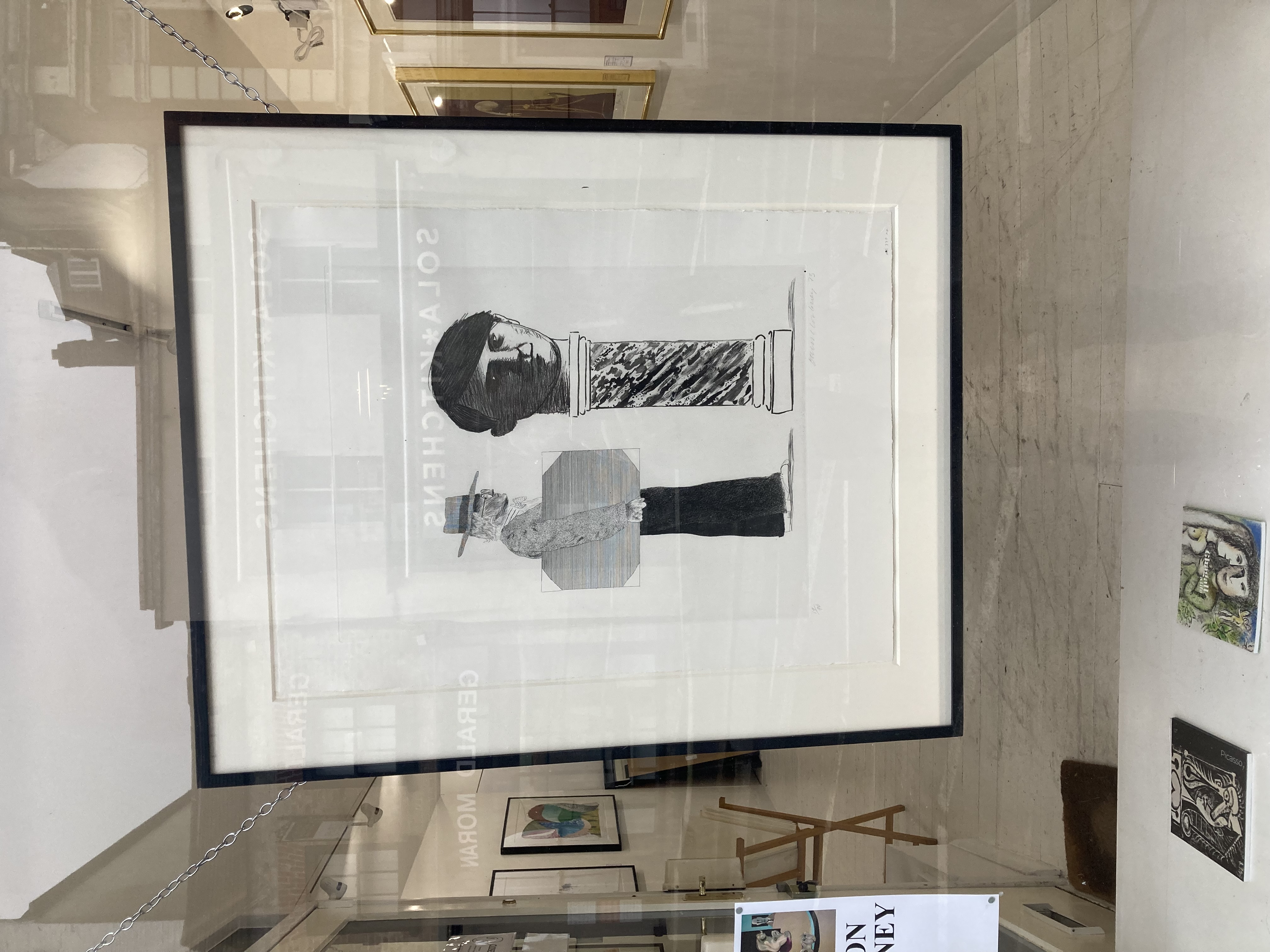 Photo in gallery window of a framed print by David Hockney, showing the artist approaching a large head of Picasso on a plinth