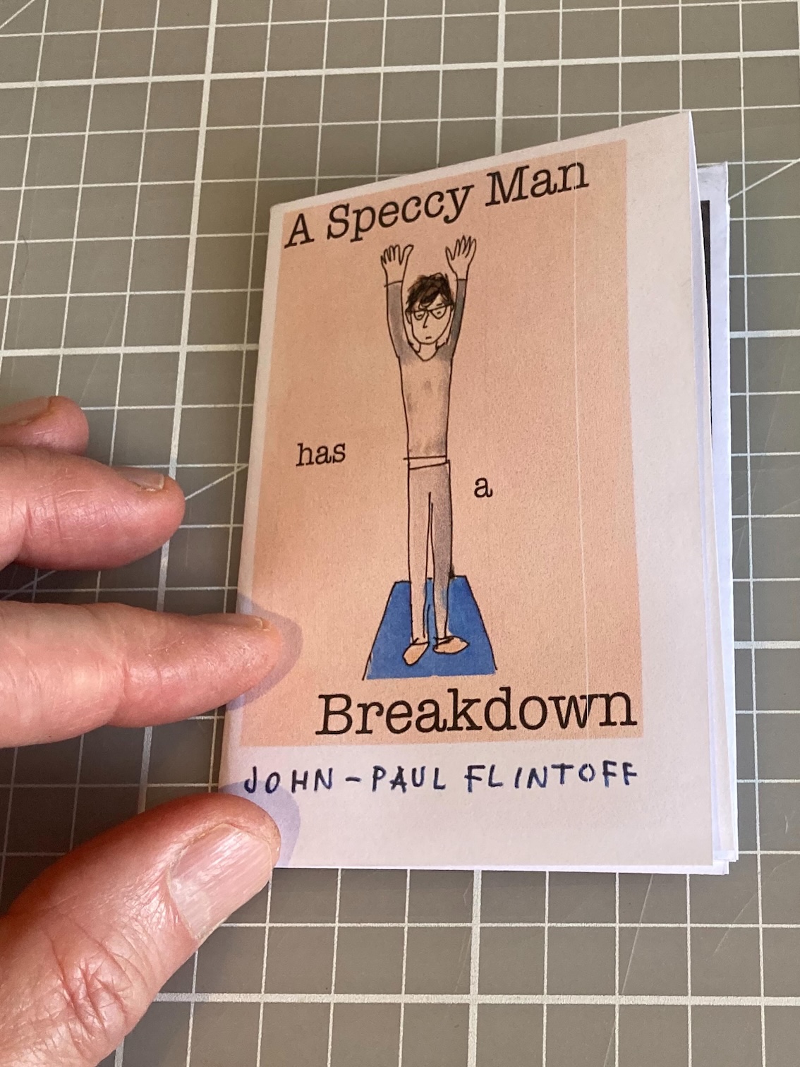 Cover of the same zine, lying down as a hand moves to open it. Cover of the same zine, lying down as a hand moves to open it.