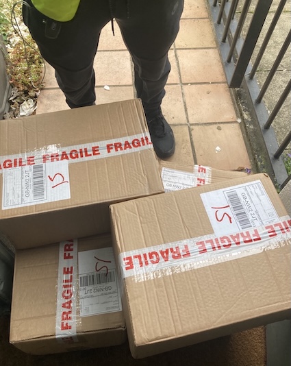 Photograph of four boxes marked FRAGILE, on a doorstep, with legs of delivery driver visible behind.