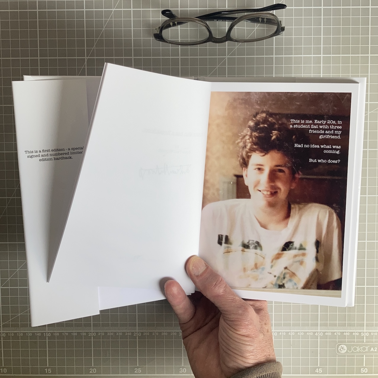 Open hardback book with the signed A5 art print displayed alongside, held in hands above a green cutting mat.