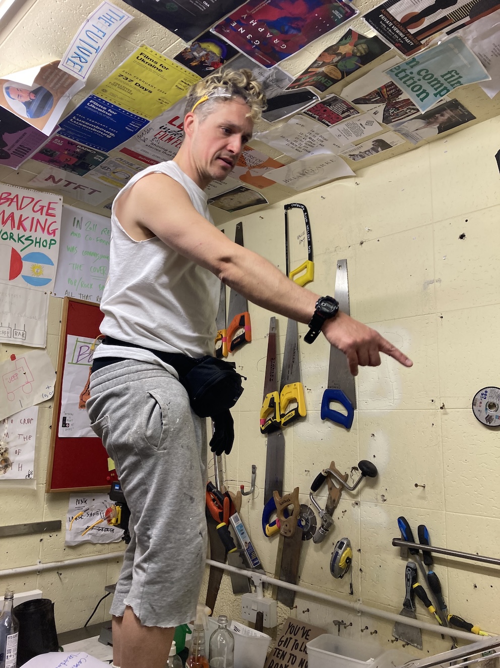 Joel Levack in a windowless workshop room at All Is Joy, in a sleeveless white vest and grey shorts, safety goggles pushed up onto his head, a black tool belt around his waist, pointing at something. The wall behind him is covered with a large collection of hand saws in different sizes and colours, along with pliers, tape measures and screwdrivers. The ceiling is covered with posters from Soho events: Films for Ukraine, badge-making workshop, film festivals, exhibition invites. Joel Levack in a windowless workshop room at All Is Joy, in a sleeveless white vest and grey shorts, safety goggles pushed up onto his head, a black tool belt around his waist, pointing at something. The wall behind him is covered with a large collection of hand saws in different sizes and colours, along with pliers, tape measures and screwdrivers. The ceiling is covered with posters from Soho events: Films for Ukraine, badge-making workshop, film festivals, exhibition invites.