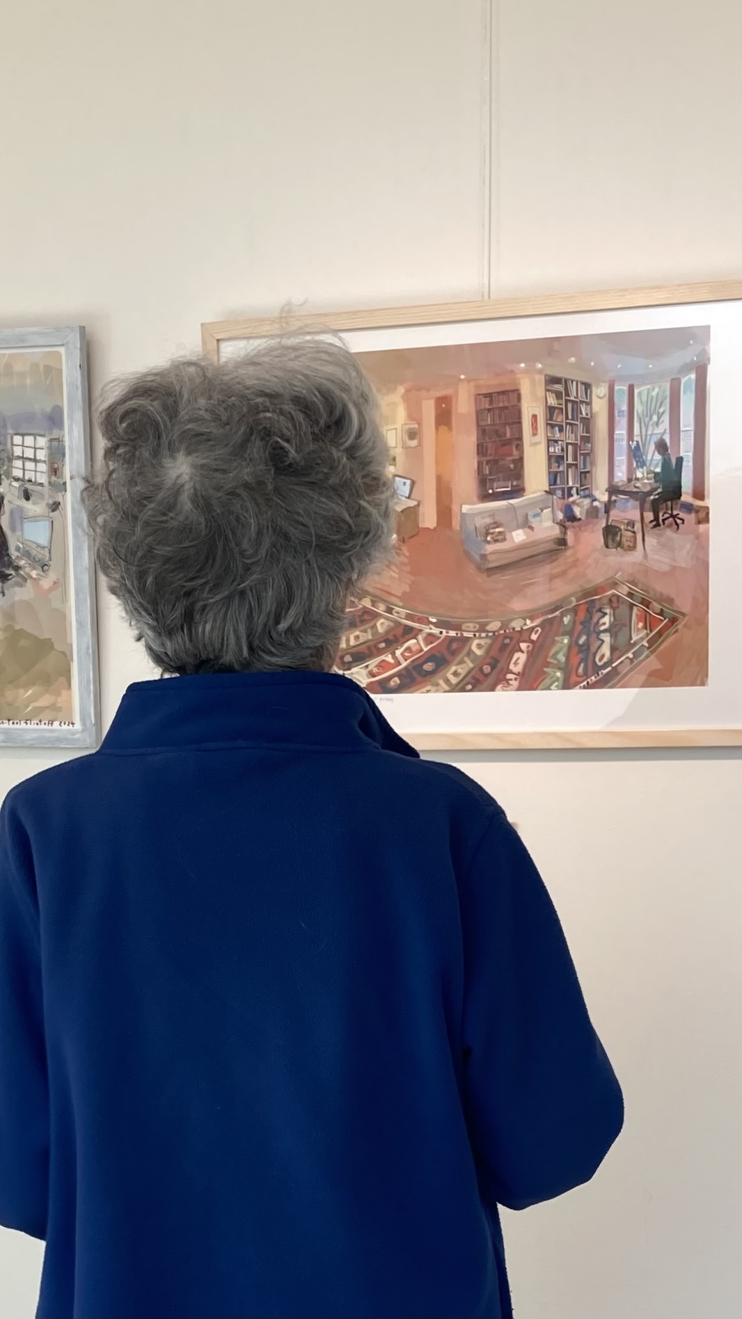 My mother, seen from behind, looks at a framed print of a book-lined living room with a patterned rug.