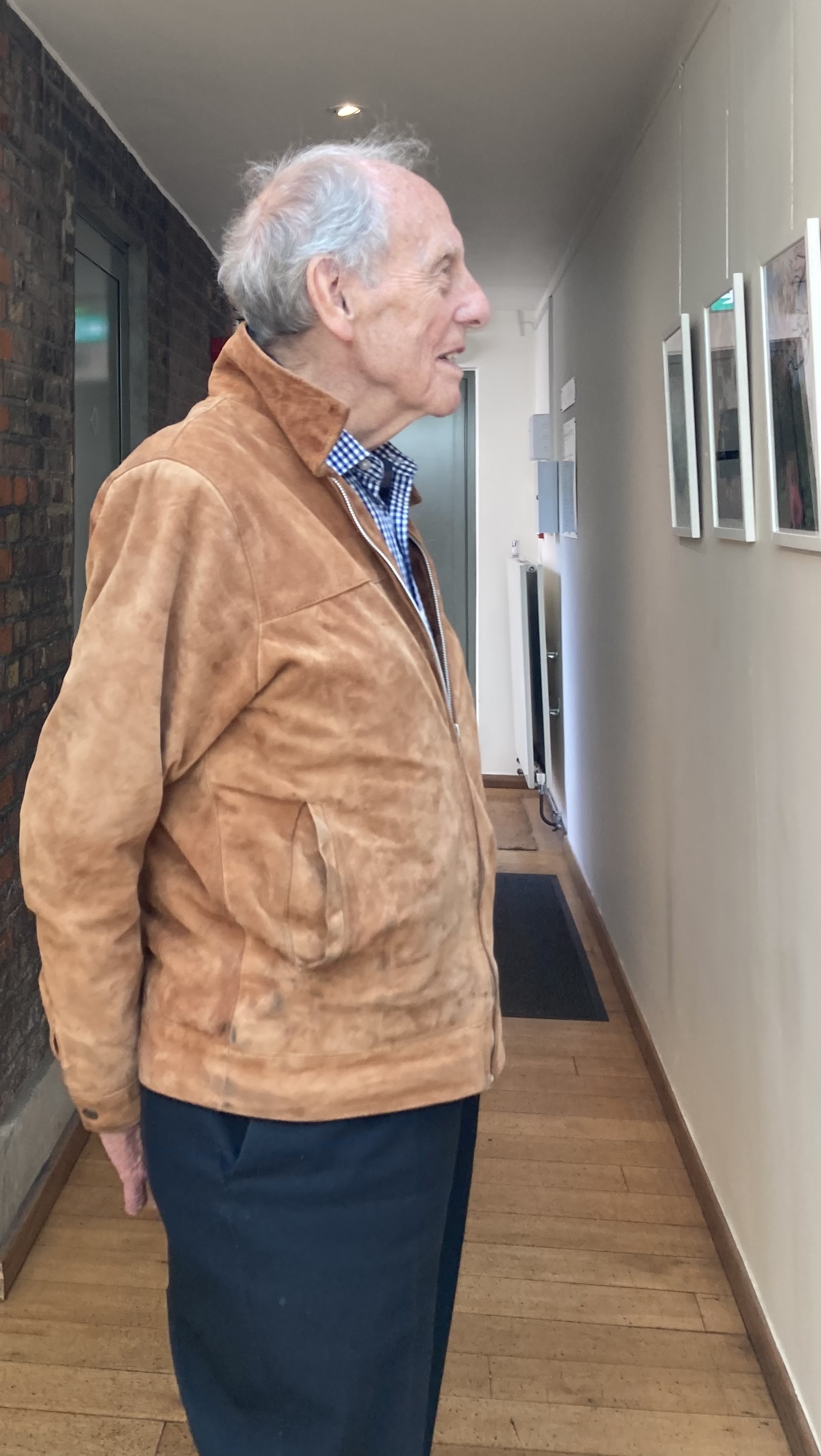 My father, in a tan suede jacket, walks along the Burgh House corridor looking at framed prints, an exit sign visible ahead.