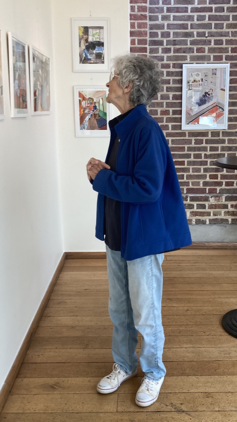 My mother, in a blue jacket, looks up at framed prints on a gallery wall beside exposed brick.