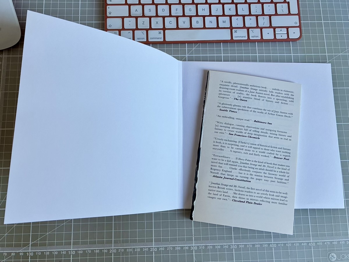 A sheet of white A3 paper cut to the height of the book, with a slim sliced section of the paperback lying on it, on a green cutting mat next to a red keyboard