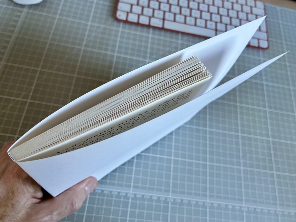 The white paper folded around the sliced book section and creased either side of the spine, held in a hand above the cutting mat