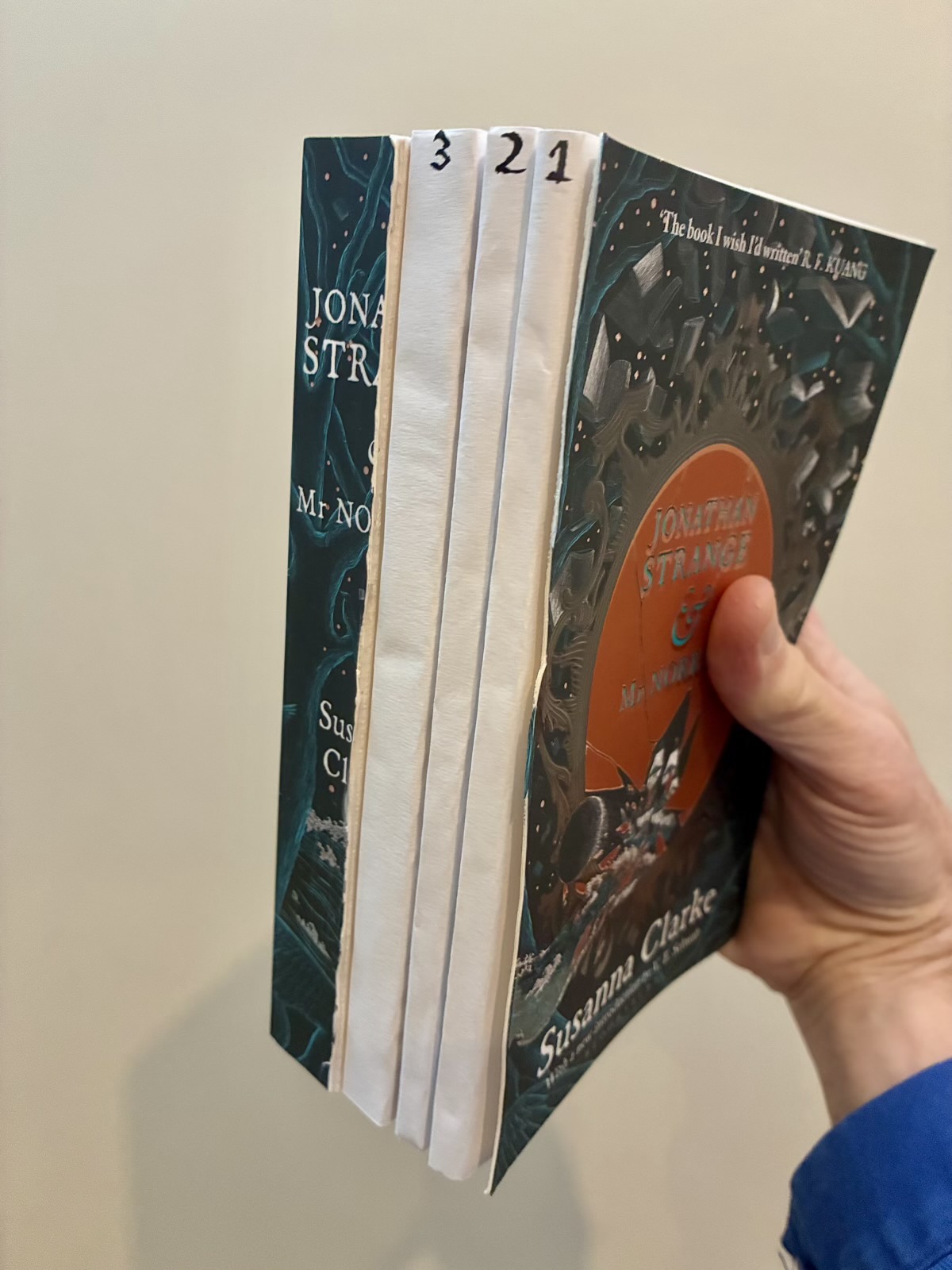 A hand holds Jonathan Strange & Mr Norrell by Susanna Clarke. Three slim sections in white paper covers, numbered 1, 2 and 3 on their spines, sit between the front cover and the rest of the book.