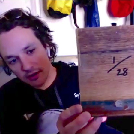 A still from a Zoom call: Joel Levack at home with dark curly hair and EarPods in, wearing a dark cycling top, holding up a small square of aged brown wood inked with "1/28". Behind him, cycling helmet and apparel on shelves. A still from a Zoom call: Joel Levack at home with dark curly hair and EarPods in, wearing a dark cycling top, holding up a small square of aged brown wood inked with "1/28". Behind him, cycling helmet and apparel on shelves.